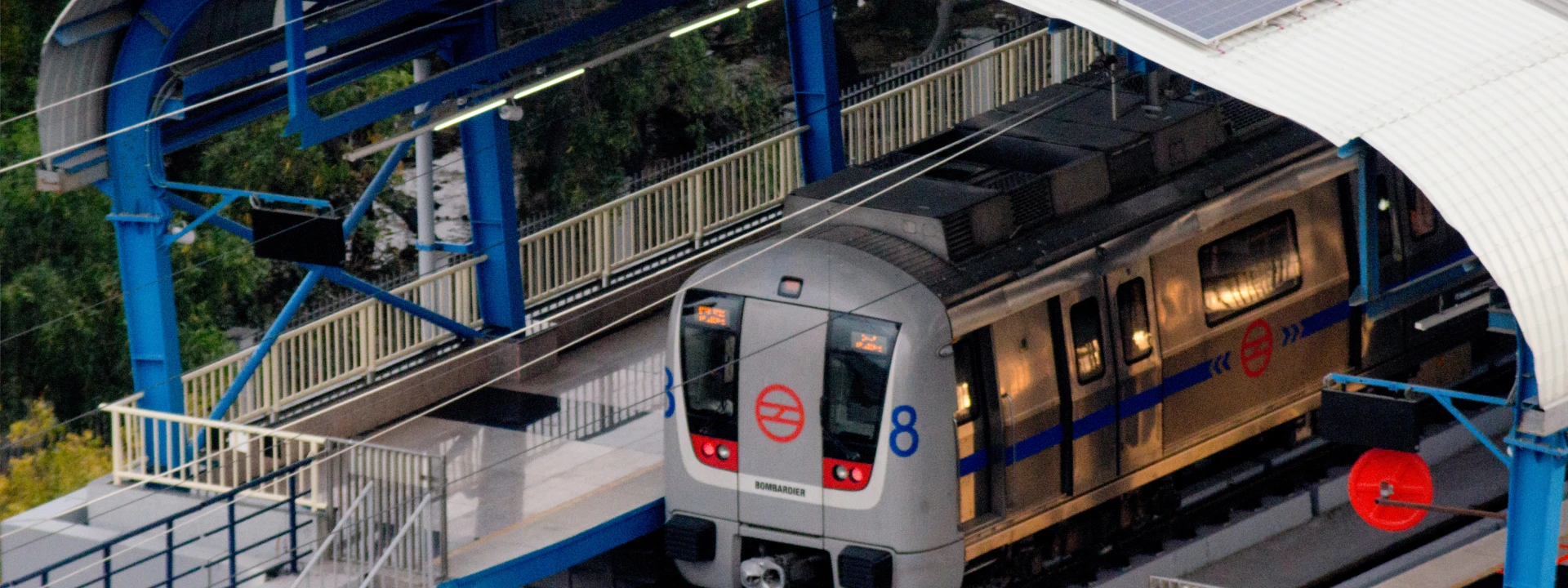 Delhi Metro train arriving at an elevated station with blue structural supports and overhead electric lines.
