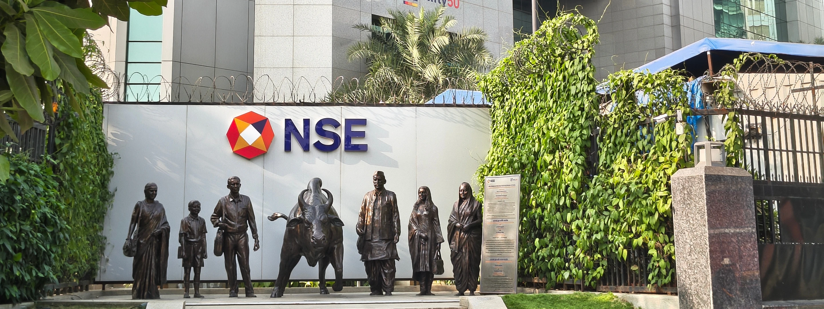 Entrance of the National Stock Exchange (NSE) building in India with logo signage and bronze sculptures representing investors and market participants, symbolizing financial markets and economic activity.
